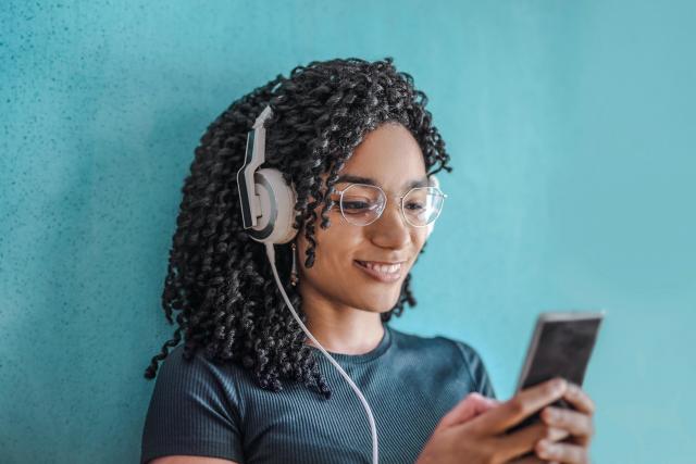 A young black woman wearing white headphones and holding an iPhone she is using to listen to audiobooks
