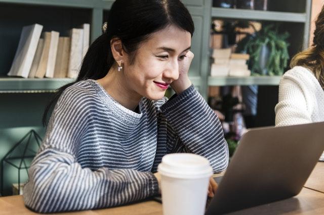 Asian woman in a blue and white striped sweater is resting her face on her hand and looking at a laptop