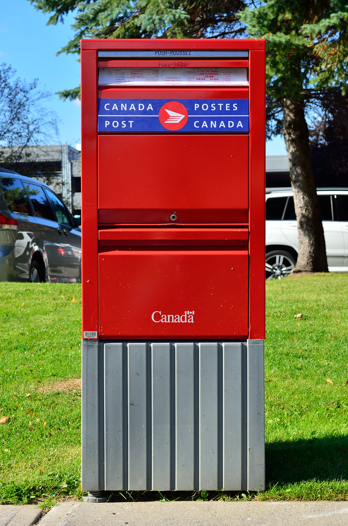 A Canada Post mailbox on a grassy lawn at the edge of a sidewalk.