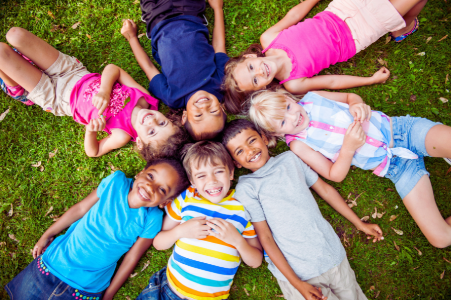Six children lie in the grass in a circle with their heads touching, smiling up at the camera.