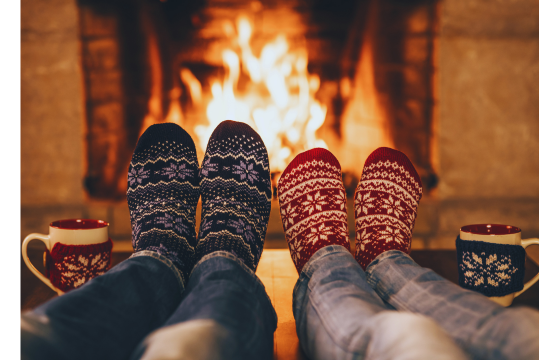 Two people visible from the knee down are warming their feet in front of a fire in a fireplace. They are wearing festive socks and have festive mugs beside them.